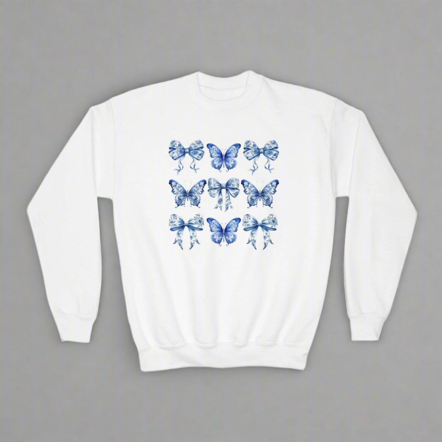 Image: This beautiful blue toile sweatshirt for girls brings cottagecore charm with butterfly and bow motifs. Sized for youth XS to XL, this cozy top matches perfectly with our toddler and women’s collections. Great for family portraits, pumpkin patches, or spring strolls. Inspired by Texas coquette fashion and the latest soft girl trends, this sweatshirt is equal parts stylish and practical, by jaecrece.