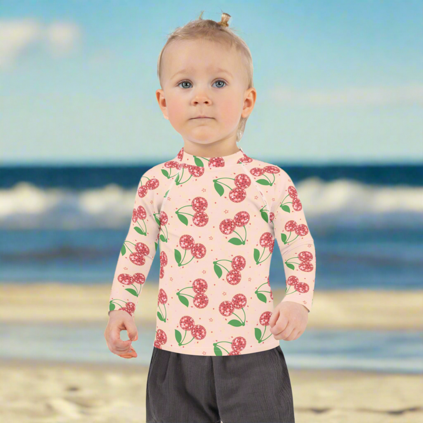 Toddler girls long sleeve rash guard featuring a bright cherry print on a soft pink base, available in sizes 2T to 7. Designed with a fitted shape and UPF 50 protection to keep kids comfortable and covered in the sun. Ideal for swimming, beachwear, and poolside play. The stretchy fabric allows easy movement while the playful design adds style. Matching mommy and me swimwear is available for coordinated family outfits.