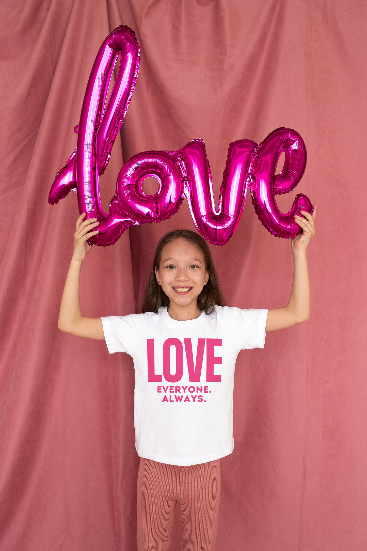 Young girl holding a hot pink balloon that spells LOVE. She is wearing a white tshirt that says "LOVE Everyone. Always" in hot pink block letters.