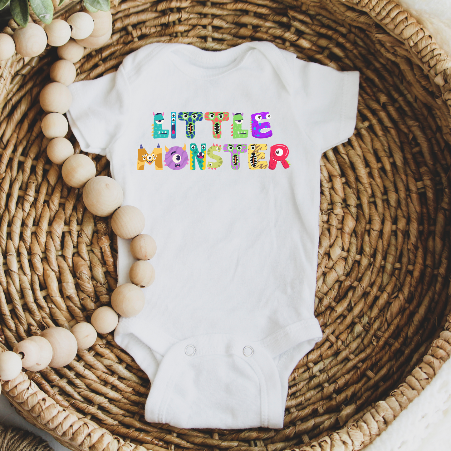 Flat lay of the white Little Monster baby bodysuit, featuring colorful cartoon monster letters spelling “little monster” across the front. Printed on a soft Bella + Canvas 100B one-piece, this breathable cotton bodysuit has an envelope neckline and three-snap leg closure for easy diaper changes. Perfect as a first Halloween outfit, fall romper, or baby shower gift, available in newborn through 24 months, and designed for everyday comfort and durability.