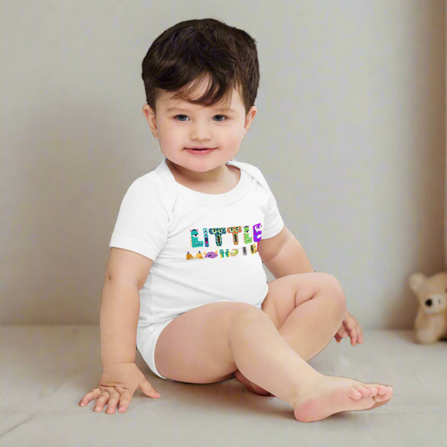 Baby boy wearing a white Little Monster bodysuit in a cozy nursery, featuring playful cartoon monster letters spelling “little monster” across the front. Printed on a soft Bella + Canvas 100B one-piece, this newborn Halloween outfit is gentle on sensitive skin and designed with an envelope neckline and three-snap closure for easy changing. Available in sizes newborn to 24 months, this fun baby shower gift doubles as a first Halloween costume alternative.