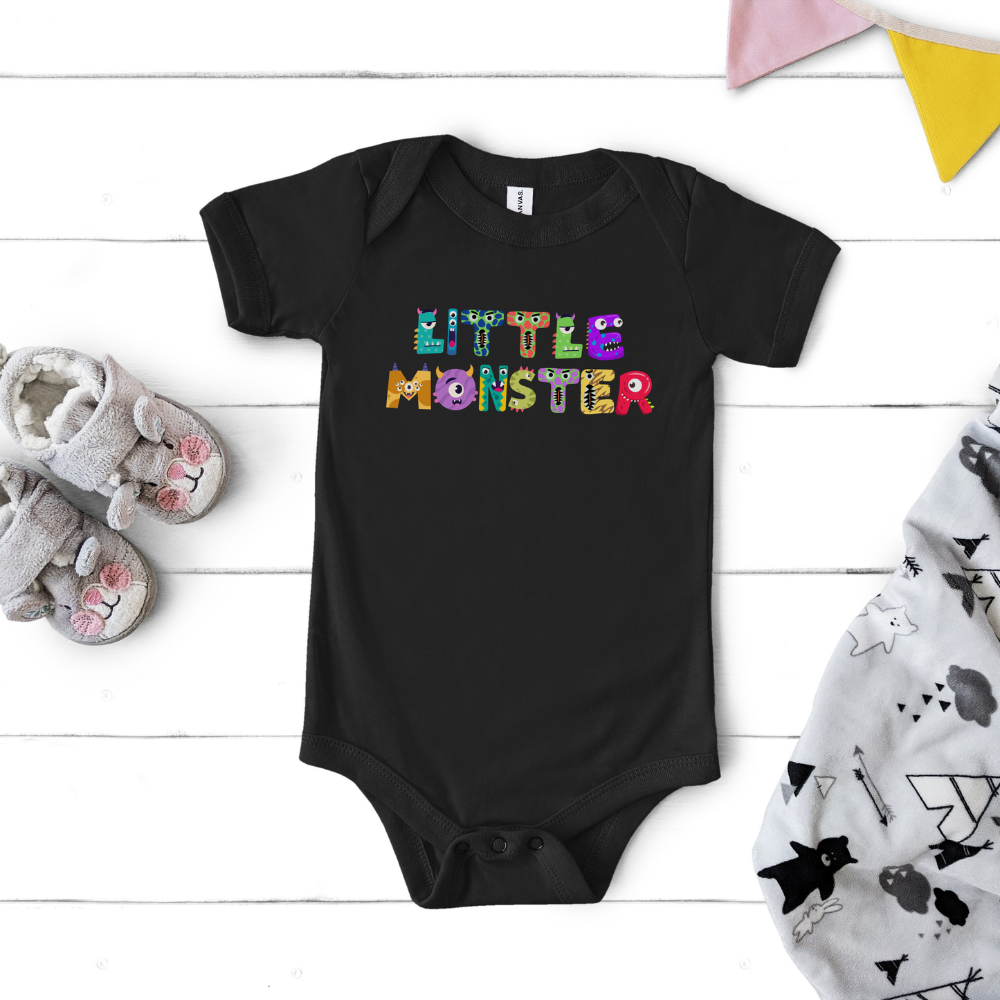 Flat lay of the black Little Monster baby bodysuit printed on a soft Bella + Canvas 100B, featuring playful cartoon monster letters spelling “little monster” across the chest. This infant one-piece is made from 100% combed ring-spun cotton with a lightweight feel, envelope neckline, and three-snap leg closure for easy diaper changes. Perfect for baby’s first Halloween, a fall baby shower gift, or everyday wear, available in newborn through 24 months.