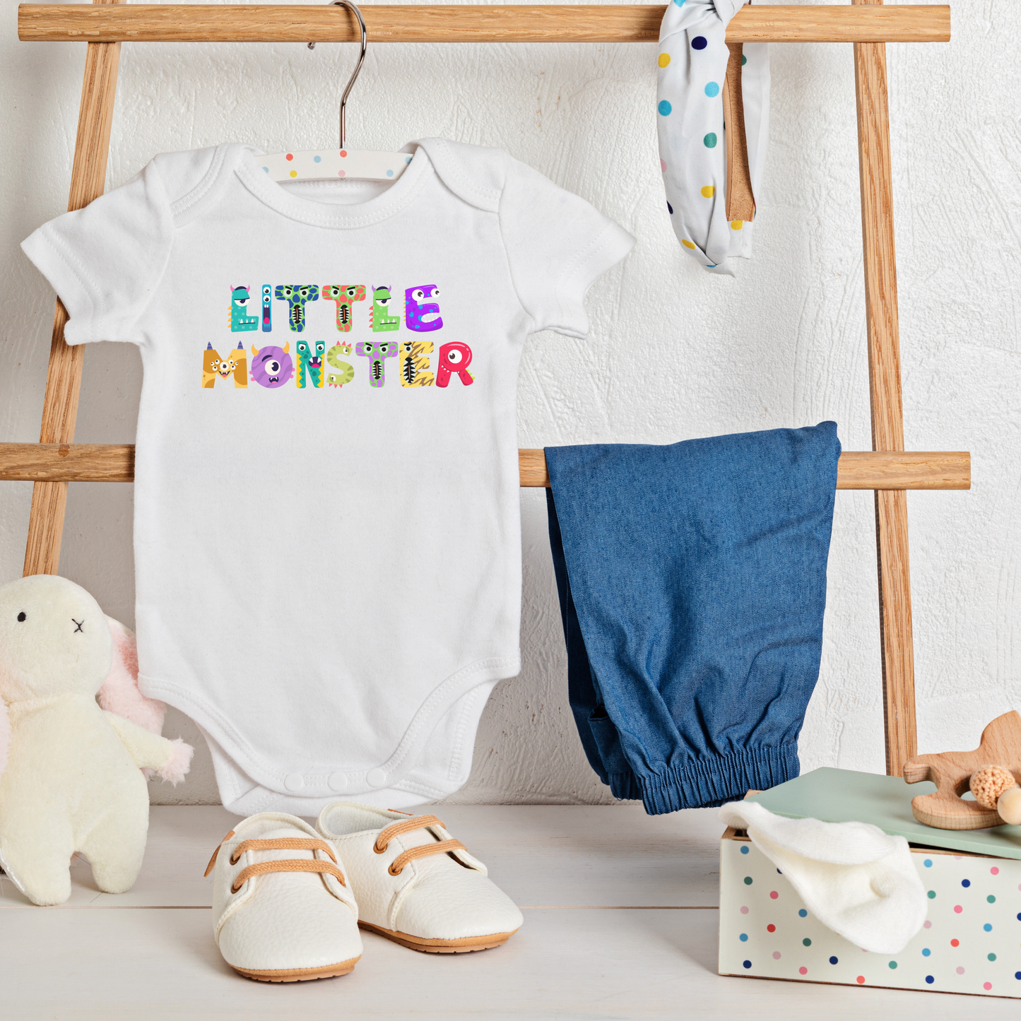 White Little Monster baby bodysuit hanging in a cozy nursery, featuring playful cartoon monster letters spelling “little monster” across the chest. Printed on a soft Bella + Canvas 100B one-piece, this infant bodysuit is crafted from 100% combed ring-spun cotton for comfort and breathability. With an envelope neckline and three-snap leg closure, it’s practical for everyday wear while doubling as a cute first Halloween outfit or thoughtful baby shower gift, newborn to 24 months.
