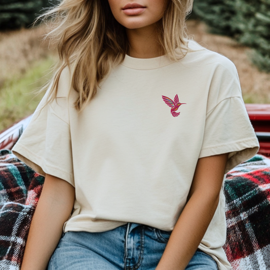 A soft cream cotton shirt with a small embroidered pink hummingbird on the chest, styled for casual, everyday wear. The subtle bird design adds a charming touch to this minimalist unisex tee, available in five classic colors and sizes XS–5XL. Ideal for birdwatching enthusiasts, garden lovers, or anyone who appreciates ethically made embroidered apparel.