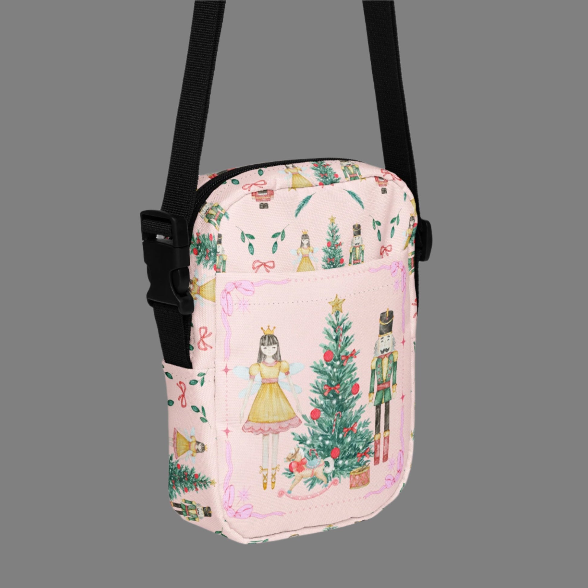Image: Pink Coquette Nutcracker Utility Crossbody bag. The front of this small mini tote has a Christmas tree, Nutcracker, Sugarplum Fairy, Rocking Horse and a holiday present The rest of the purse is covered in a pattern of ribbons, garland, and Nutcrackers. May be worn across the waist/ hips, across the chest or as a mini backpack, by jaecrece