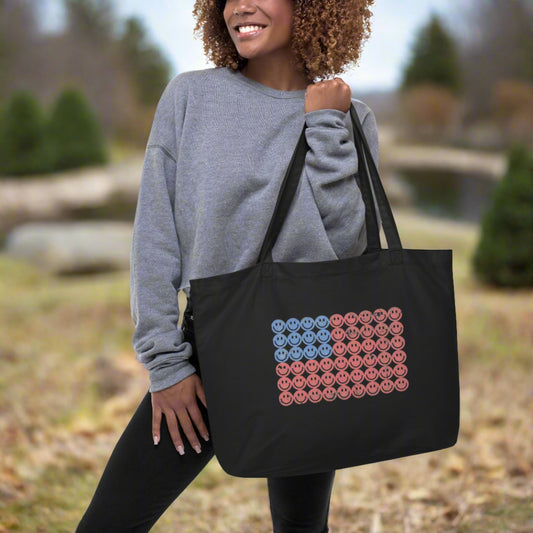 Large 100% organic cotton tote bag in black featuring a vibrant American flag graphic made from red, white, and blue retro smiley faces. This oversized eco-friendly tote is perfect for summer, Fourth of July events, farmers markets, beach days, and patriotic gifting. Durable, reusable, and stylish for everyday errands with a nostalgic twist.