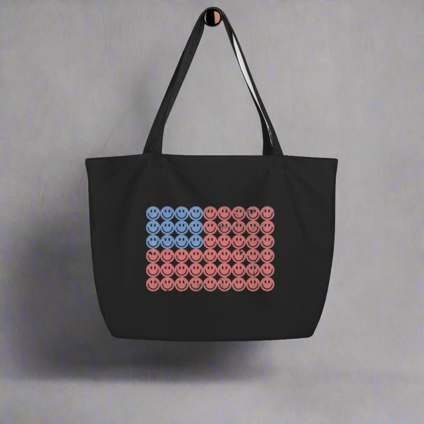 Black organic cotton tote bag featuring a large American flag design made from red, white, and blue retro smiley faces. This oversized patriotic tote is perfect for the Fourth of July, beach trips, shopping, or summer festivals. A trendy and sustainable option for vintage lovers and eco-conscious shoppers alike. Keywords: American flag tote, retro smiley face bag, large organic cotton tote, patriotic summer bag, reusable canvas tote.