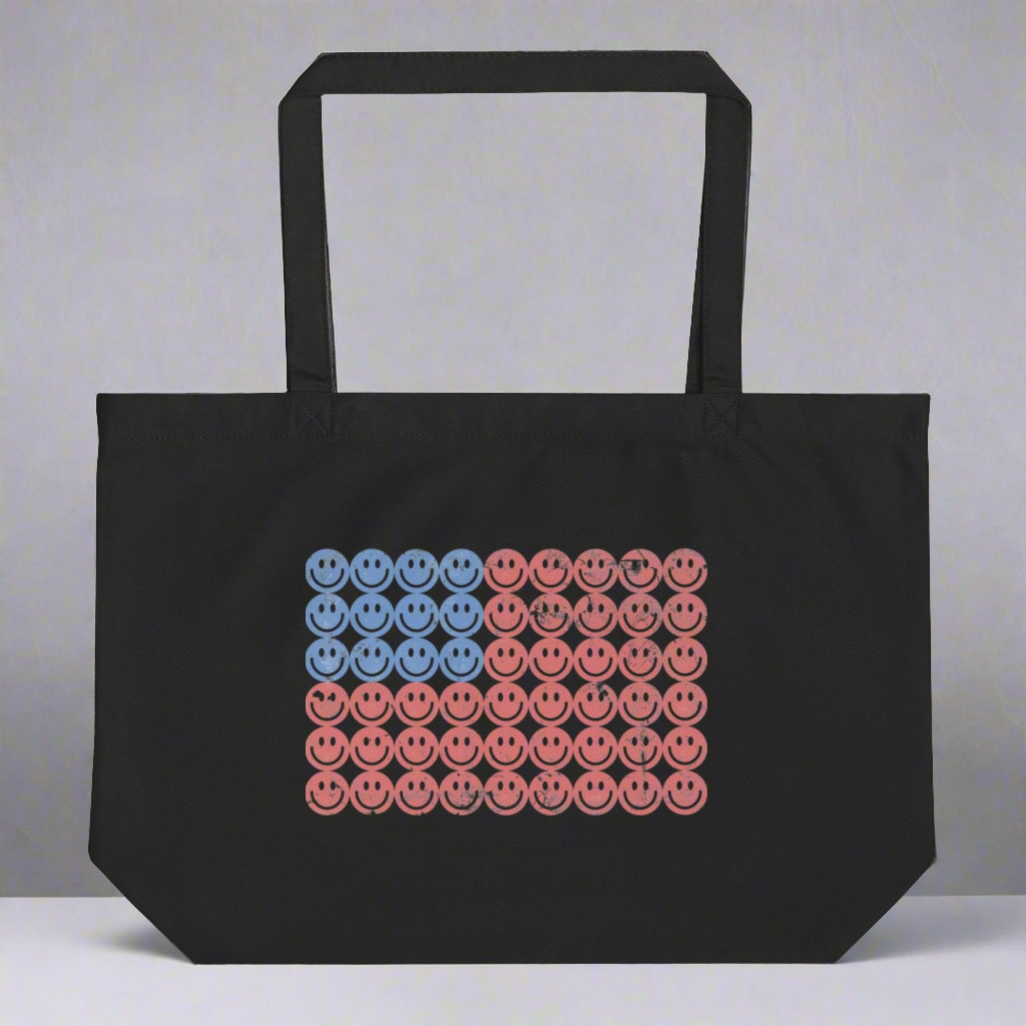 Black 100% organic cotton tote bag with a whimsical American flag print made of colorful smiley faces in red, white, and blue. This large, reusable tote is perfect for summer festivals, beach days, and showing off your patriotic side in vintage style. Durable and fun, it’s a great accessory for anyone who loves smiley face graphics and sustainable fashion. Keywords: patriotic tote, retro smiley tote, Fourth of July flag bag, organic cotton shopping bag.
