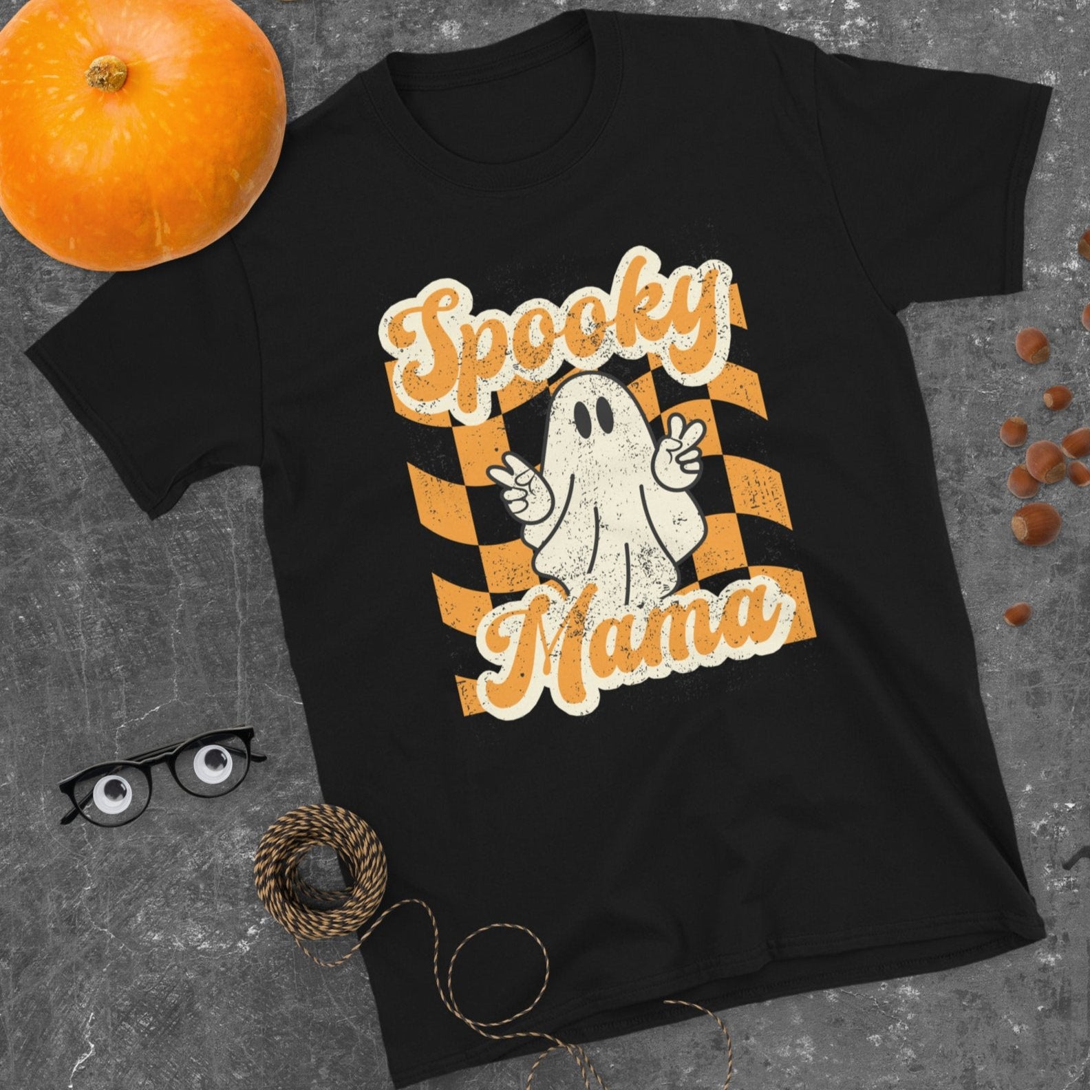 Spooky Mama Halloween Tshirt, featuring a orange checkerboard design with a white ghost giving the peace sign, and the text "Spooky Mama" in orange and white cursive bubble font, by jaecrece.com