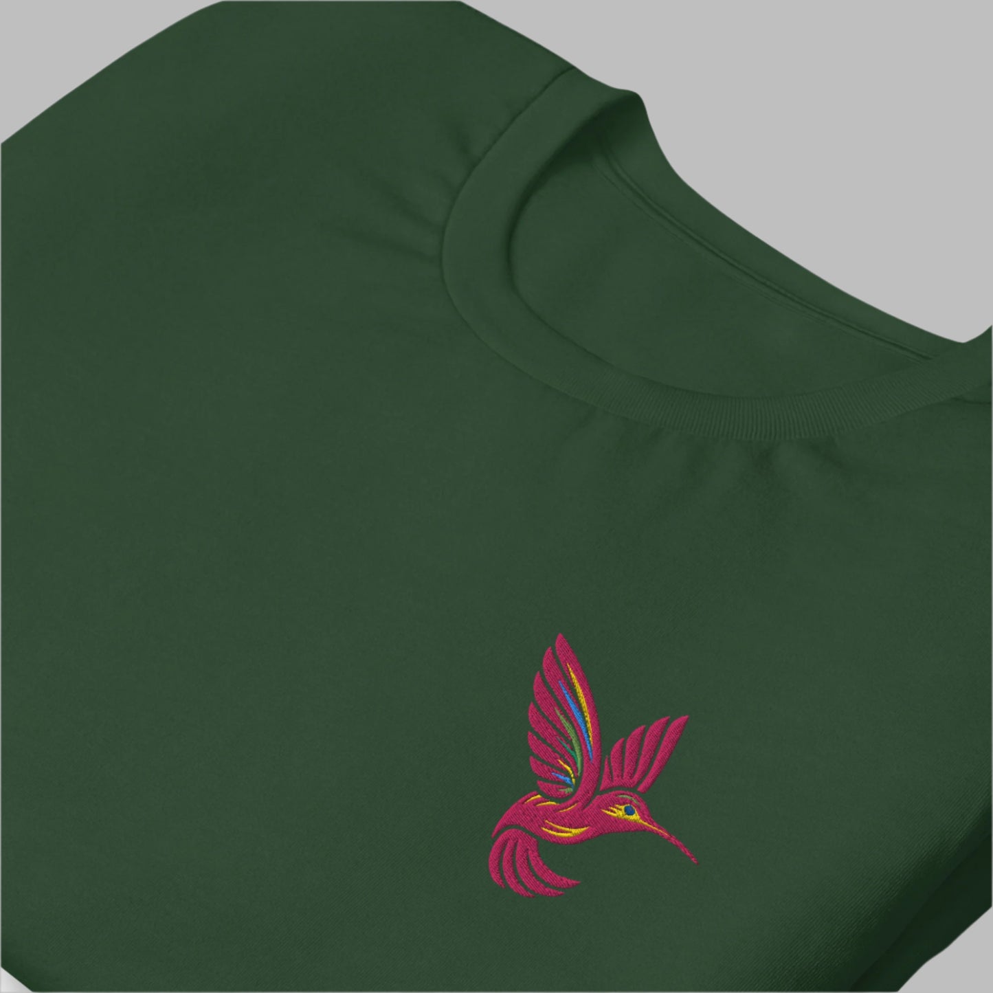A soft cotton unisex t-shirt in forest green, featuring a bright pink hummingbird embroidered on the left chest. This minimalist nature-inspired shirt is available in sizes XS–5XL and makes a great gift for bird lovers, gardeners, and fans of embroidered details. A sustainable, pre-shrunk tee perfect for casual wear, spring fashion, or cottagecore style lovers looking for something unique and charming.