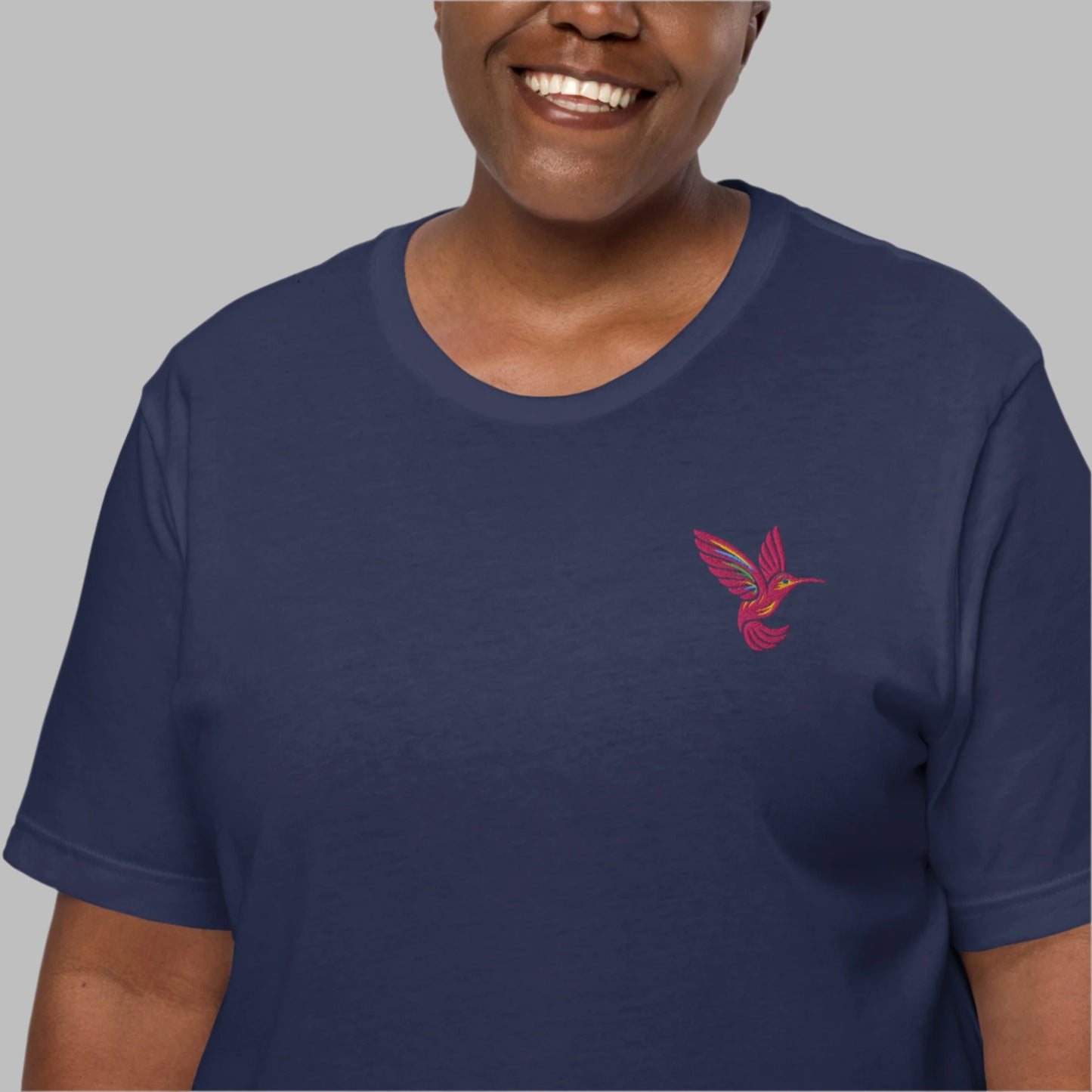 A woman wears the navy version of the Hummingbird Embroidered Shirt, showcasing a bright pink hummingbird stitched delicately over the left chest. Made from soft, breathable cotton, this nature-inspired shirt blends minimalist embroidered detail with everyday comfort. Available in sizes XS–5XL, this slow fashion piece is ideal for bird lovers, spring outfits, cottagecore fashion, and sustainable gift ideas.