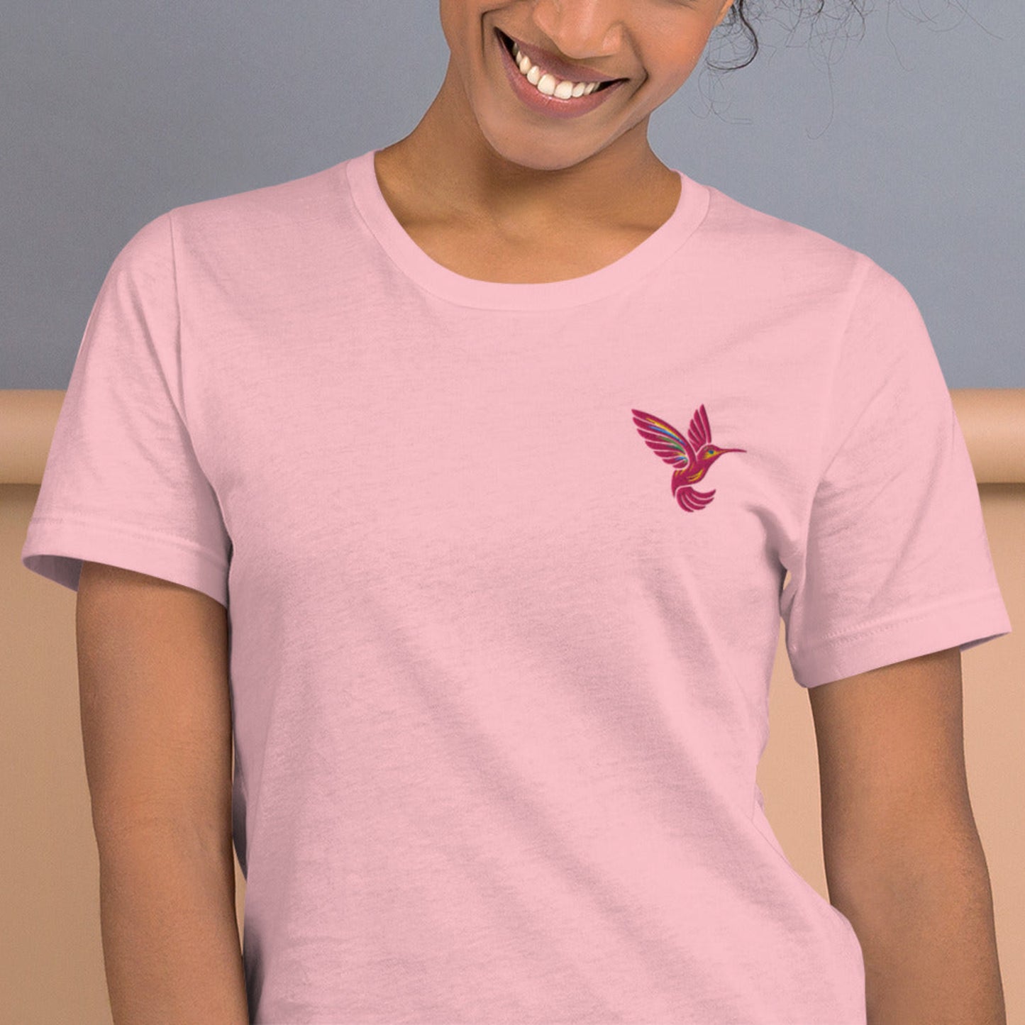 Front view of a pastel pink t-shirt featuring a single bright pink hummingbird embroidered over the left breast. This shirt blends casual comfort with whimsical nature-inspired design, available in sizes XS through 5XL. Ideal for spring outfits, cottagecore aesthetics, or gifting to women who love hummingbirds, embroidery, or eco-conscious fashion made on demand.