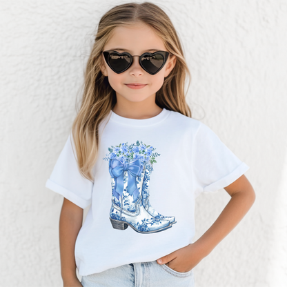 Image: A dreamy blend of western charm and girly flair, this youth-sized shirt showcases a pair of cowboy boots adorned with a blue bow and wildflowers. Designed for girls who love country fashion, it brings coastal cowgirl style into everyday wear. Available in sizes S to XL, this tee is cozy and stylish—ideal for birthdays, holidays, or any outfit upgrade, by jaecrece.
