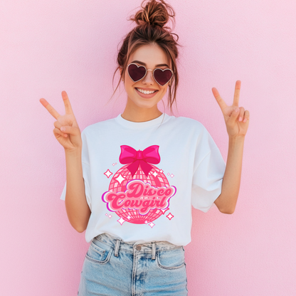 Disco Cowgirl women's graphic tee in Ivory features a large pink disco ball, hot pink bow, and starry background with retro script reading "Disco Cowgirl." This Comfort Colors 1717 t-shirt brings coquette western style to your next Nashville trip, rodeo, or honky-tonk party. Boutique-inspired, perfect for bachelorette celebrations or matching girls' night out looks. Available in sizes S–4XL.

