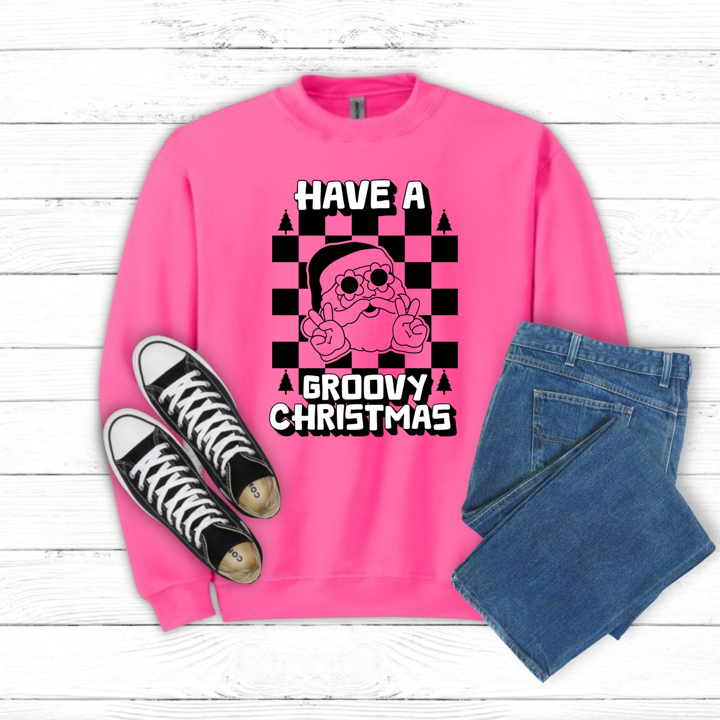 Image: retro Santa christmas sweatshirt available in pink and bright green. The text reads Have a groovy Christmas, with a black checkerboard background, and a vintage Santa wearing daisy sunglasses and giving the peace sign. The perfect gift for those who love a funky vintage aesthetic, by jaecrece