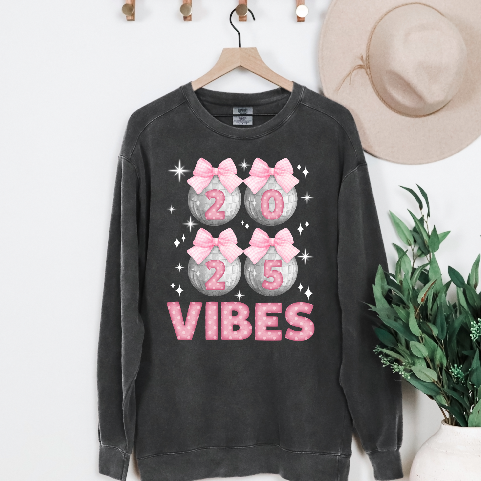 Image: Happy New Years Eve Sweatshirt is available in four colors- denim, graphite, white and chalky mint. There are 4 silver disco balls, with  pink gingham ribbon and 2025 on each. VIBES is in pink coquette polka dot block letters. by jaecrece