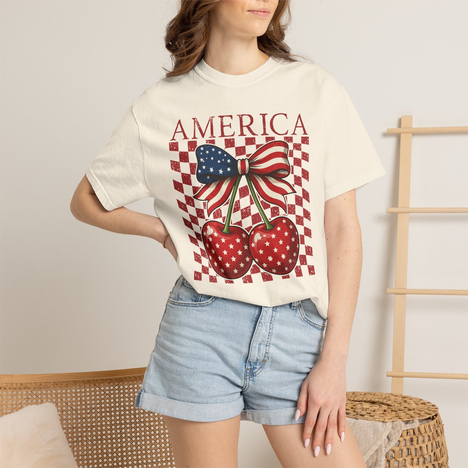 Woman wearing Ivory Comfort Colors America Cherry Bow shirt styled as a casual summer outfit. Soft ivory garment dyed tee features bold America lettering above two glossy red cherries with white star details tied together by a stars and stripes bow. Distressed checkerboard background adds retro vintage charm. Relaxed unisex fit pairs easily with denim shorts or jeans. Cute patriotic women’s shirt for July fourth, fireworks, parades, summer vacations, brunch, concerts, and everyday Americana style.