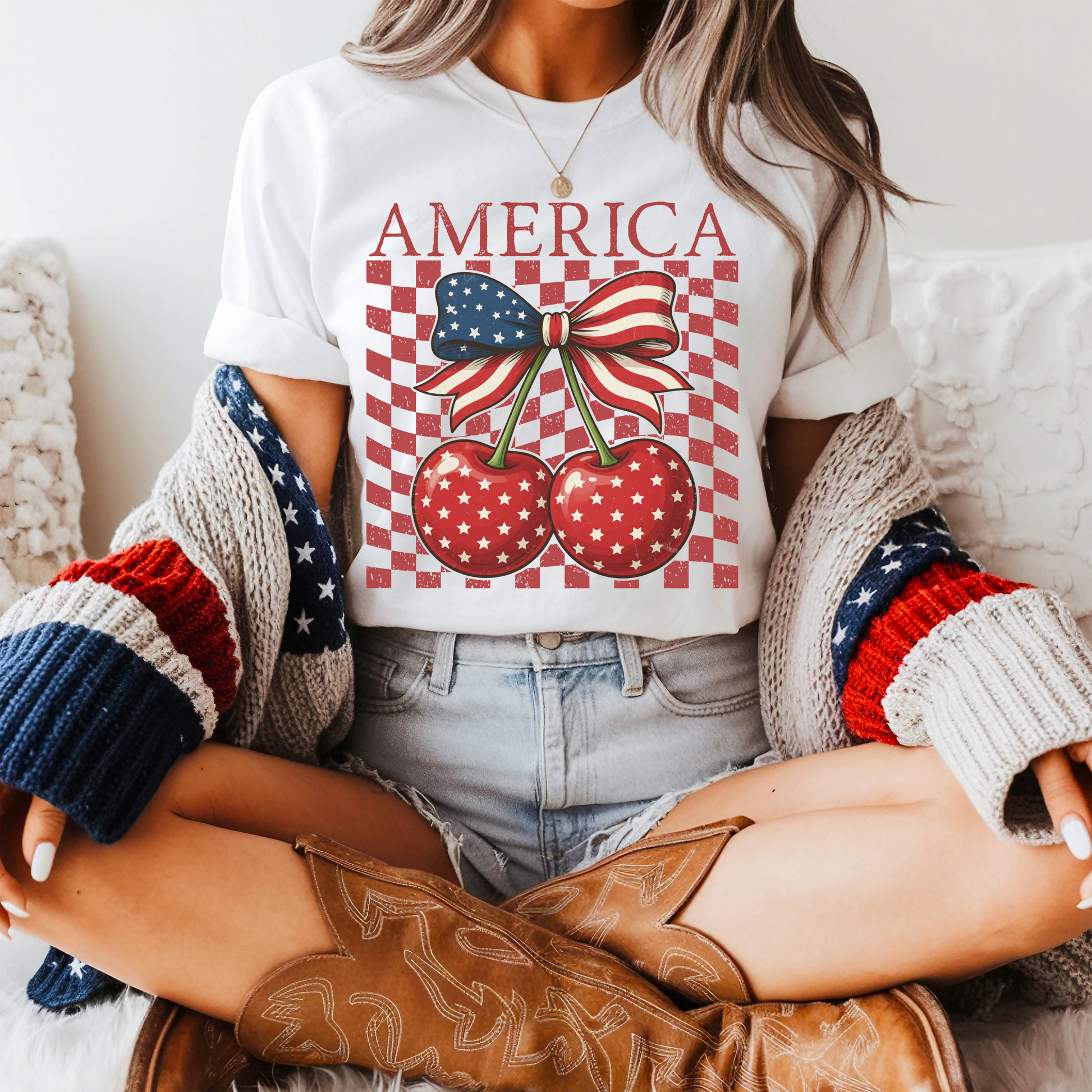 Comfort Colors garment dyed shirt with vintage America design featuring patriotic cherries and bow artwork. Large distressed letters spell America above two red star printed cherries tied together by a stars and stripes ribbon. Red checkerboard background adds trendy retro racer style. Relaxed unisex cotton tee available in multiple colors including white ivory chambray navy and blossom. Great summer holiday shirt for travel, cookouts, fireworks, and casual patriotic fashion.