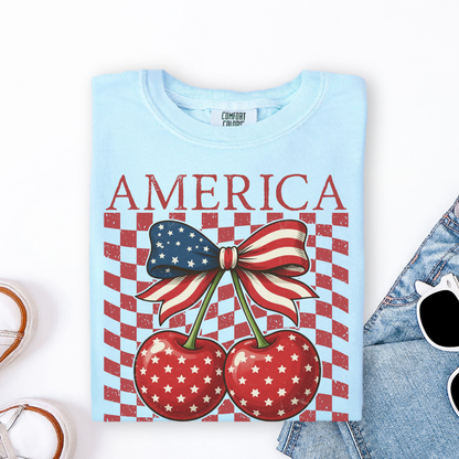 Girls Comfort Colors youth shirt in chambray featuring bold America lettering above two glossy red cherries with white star details tied together by a stars and stripes bow. Distressed checkerboard background adds vintage patriotic style. Soft garment dyed cotton tee with classic youth fit made for comfort and play. Cute girls patriotic shirt for July fourth parades, fireworks, summer vacations, school spirit days, family photos, and matching mommy and me outfits with the adult version.