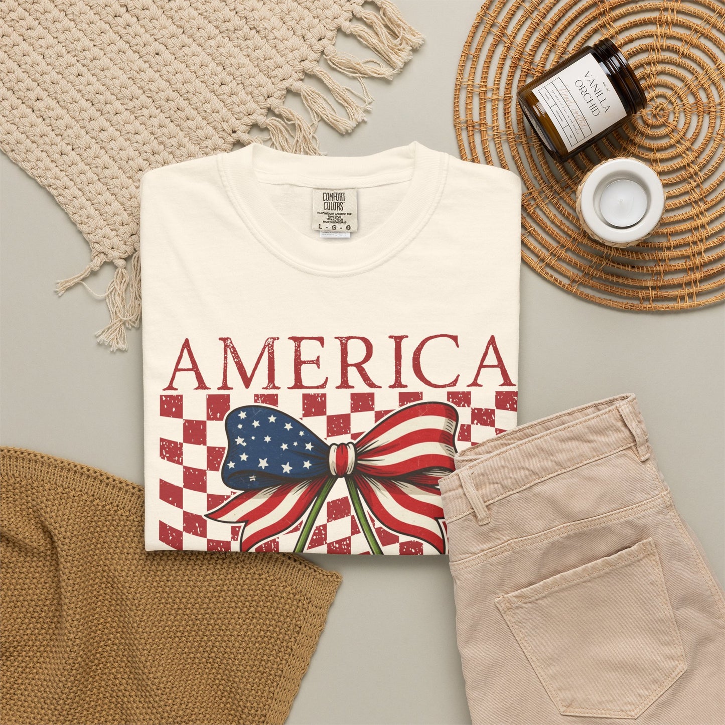 Ivory Comfort Colors patriotic graphic tee featuring America text above a retro cherry bow design. Two bright red cherries with white star details hang beneath a bow patterned with blue stars and red stripes. Distressed checkerboard background adds vintage race day style. Garment dyed cotton shirt with soft lived in feel and relaxed fit. Great for women who love coquette Americana fashion, Fourth of July celebrations, concerts, beach trips, and summer weekends.