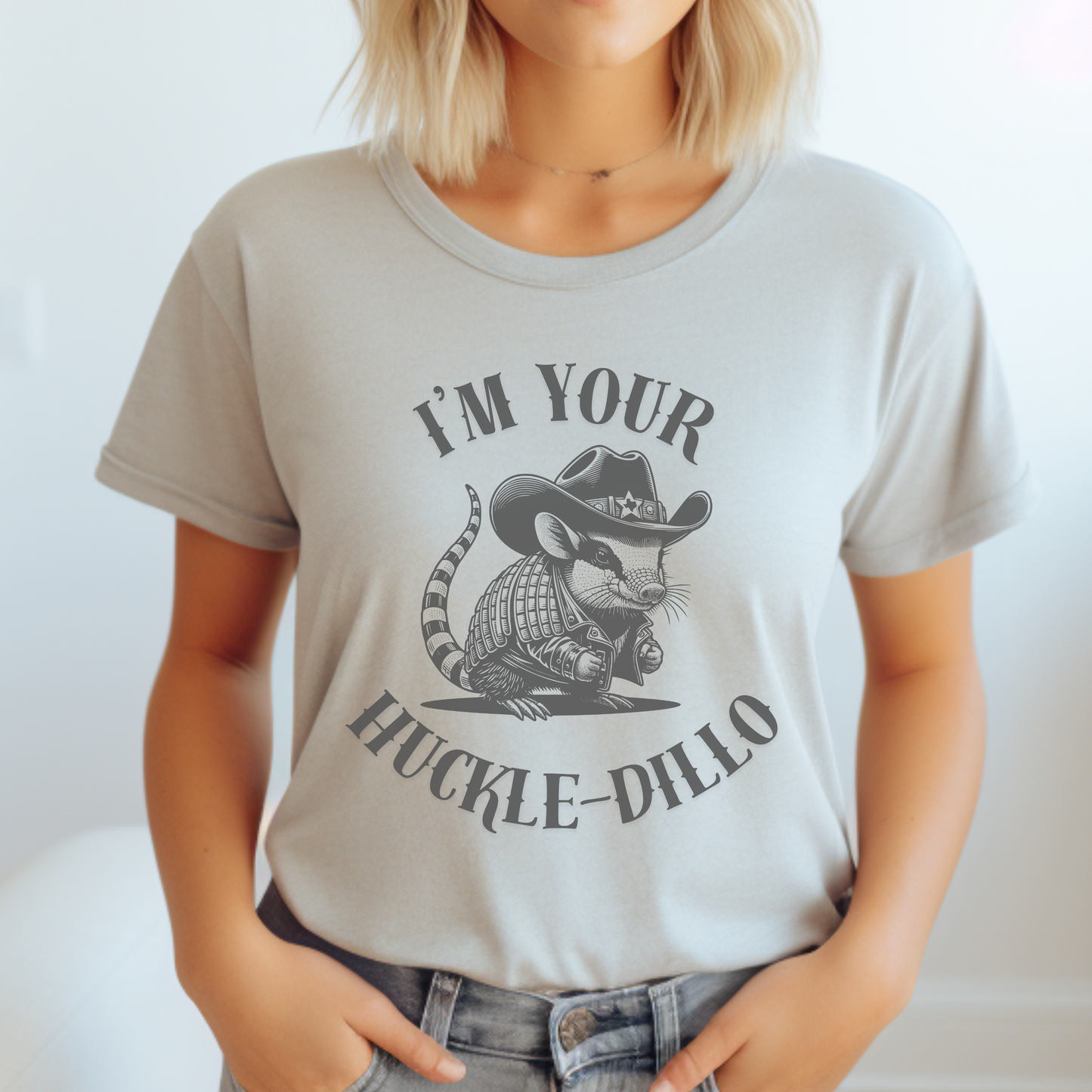 Image: A unisex T-shirt featuring a humorous dark grey illustration of an armadillo dressed as a sheriff, complete with a cowboy hat and jacket. The hat has a small Texas emblem on the brim, adding a fun southern touch. This unique design is perfect for animal lovers, western fashion fans, and anyone who appreciates quirky Texas-themed apparel. A great gift for country style enthusiasts or armadillo fans looking for a lighthearted and stylish shirt, by jaecrece.