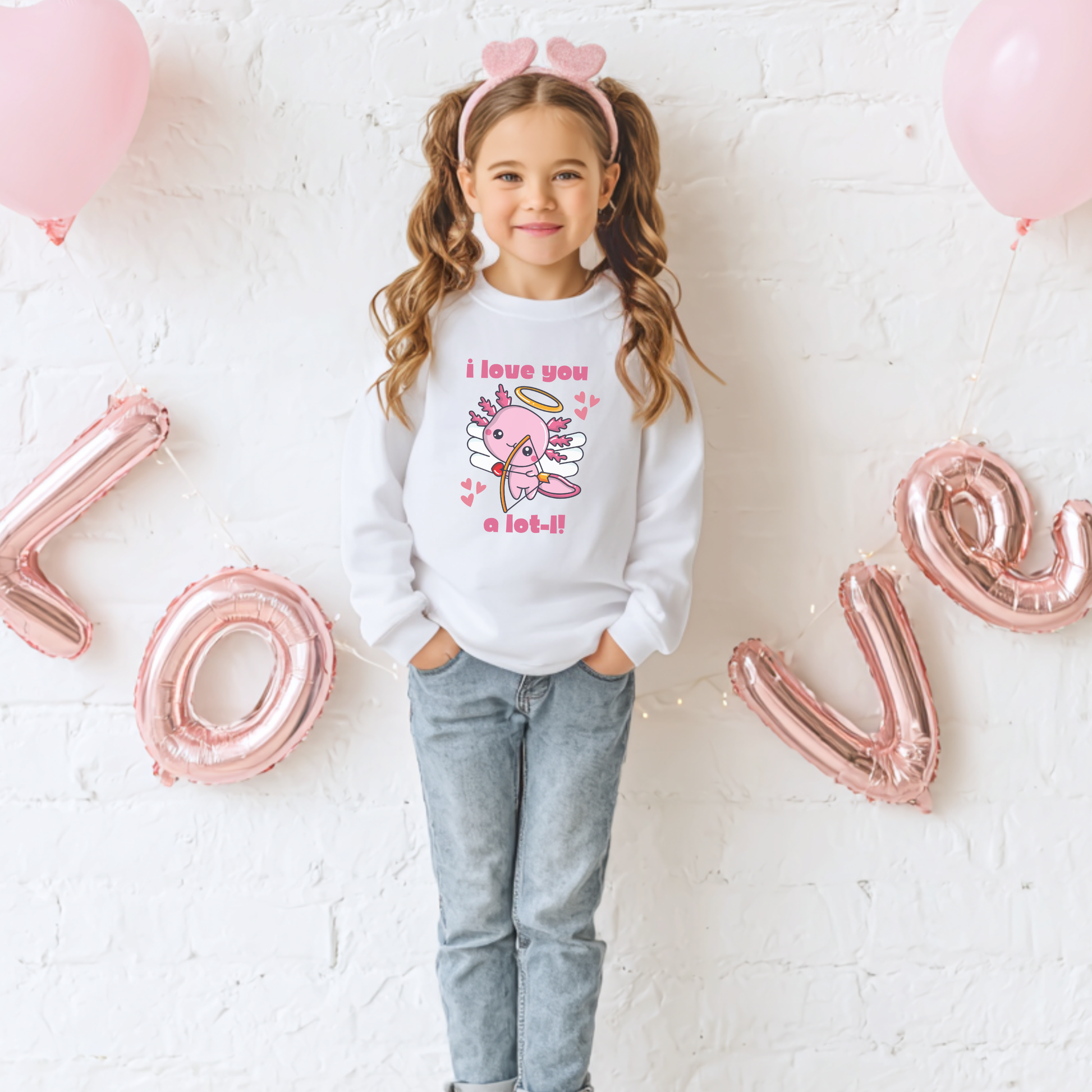 A young girl wearing a white youth Valentine sweatshirt featuring a cute axolotl cupid design with wings, halo, heart accents, and the phrase I love you a lot l. Styled against a festive Valentine themed background with soft pink decor, this cozy crewneck is perfect for school Valentine parties, classroom celebrations, and cheerful seasonal outfits for kids who love animals and playful designs.
