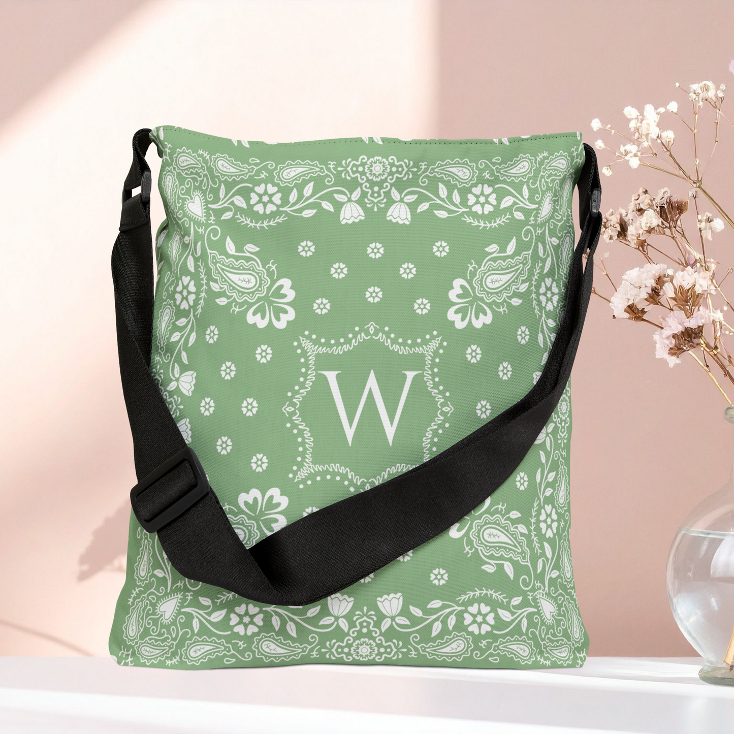 A sage green bandana print shoulder bag with a black adjustable strap, featuring a white monogram letter 'W' in the center, displayed on a white surface against a soft pink background.
