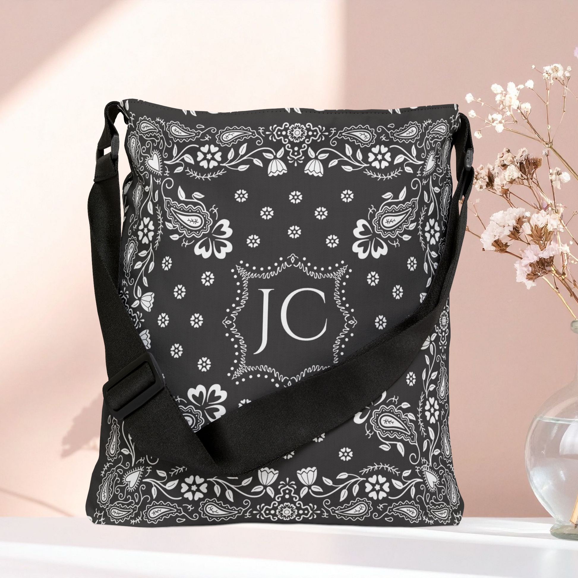 A black shoulder bag with a white paisley bandana print and a central monogram 'JC', displayed on a white surface against a soft pink background with dried flowers in a glass vase.