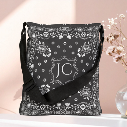 A black shoulder bag with a white paisley bandana print and a central monogram 'JC', displayed on a white surface against a soft pink background with dried flowers in a glass vase.