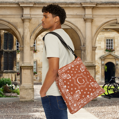 A man wearing a white t-shirt and blue jeans stands in a stone courtyard, carrying a burnt orange colored bandana tote bag with a white paisley print and a monogram letter 'G' on the front.