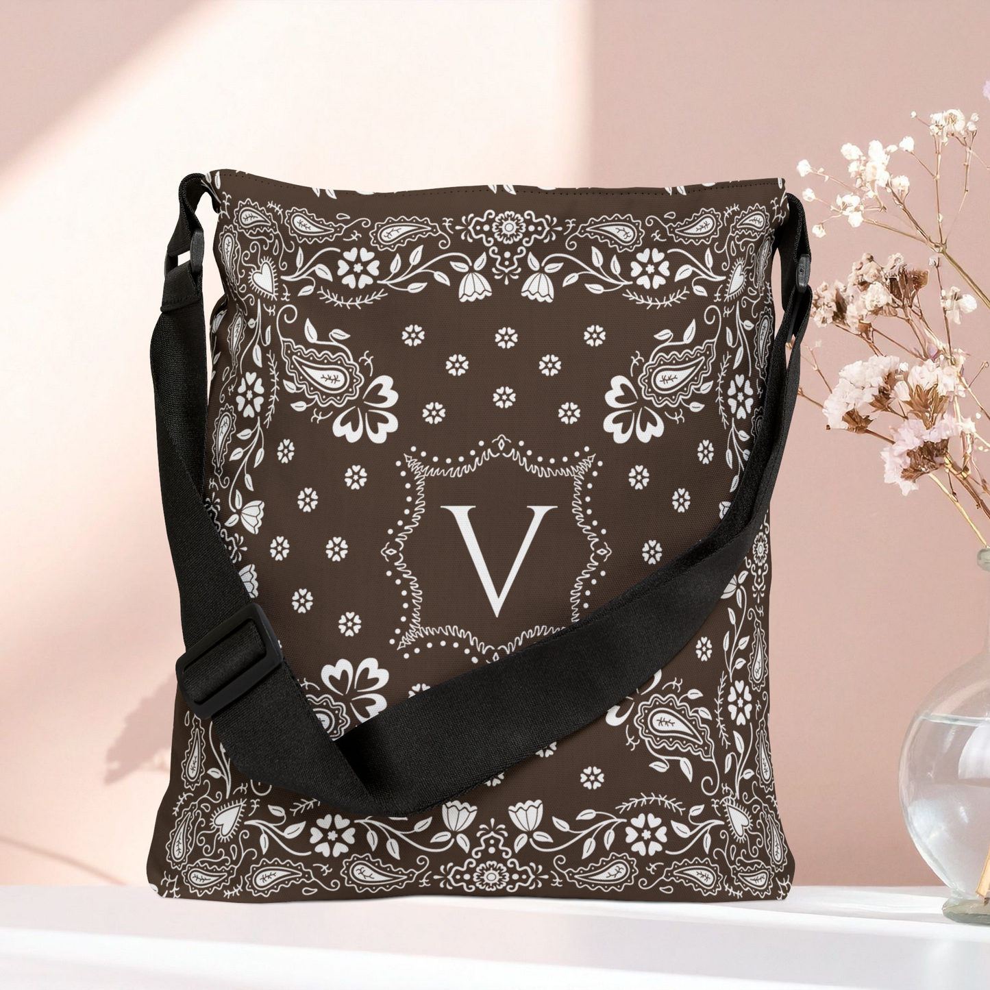 A brown bandana patterned shoulder bag with a black strap, featuring a white monogram letter 'V' in the center, displayed on a white surface against a soft pink background with dried flowers in a vase.