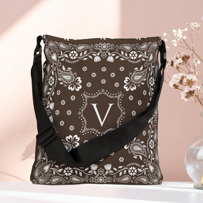 A brown bandana patterned shoulder bag with a black strap, featuring a white monogram letter 'V' in the center, displayed on a white surface against a soft pink background with dried flowers in a vase.