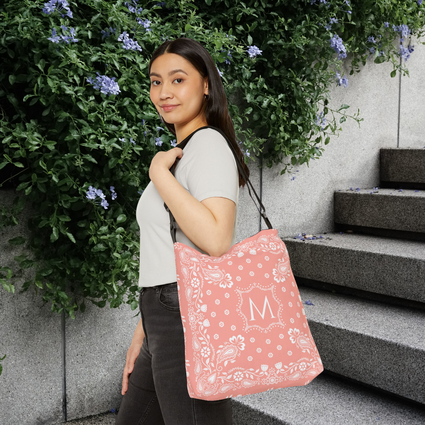 A woman wearing a white t-shirt and dark jeans carries a pink bandana patterned tote bag with a white monogram letter 'M' on a gray stone staircase next to a green leafy wall.