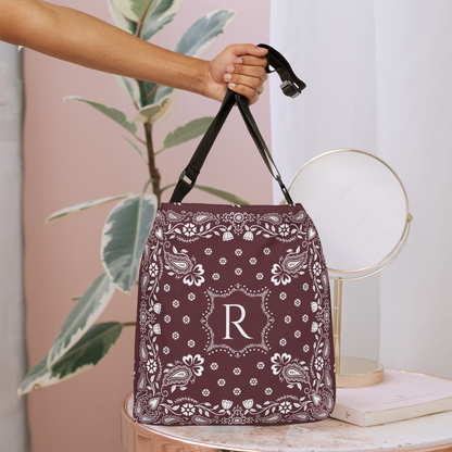 A hand holds a burgundy bandana-patterned bucket bag with black handles, featuring a white monogram letter 'R' in the center, set against a soft pink and white background with a plant and a small mirror.