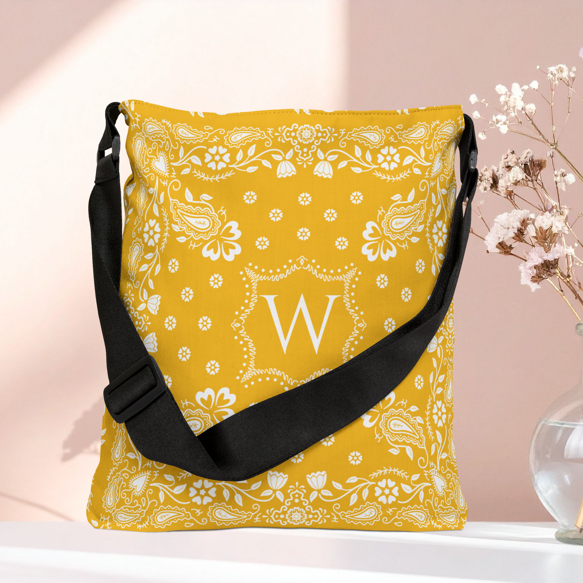 A yellow bandana patterned crossbody bag with a black strap, featuring a white monogram letter 'W' in the center, displayed on a white surface against a soft pink background with dried flowers in a vase.