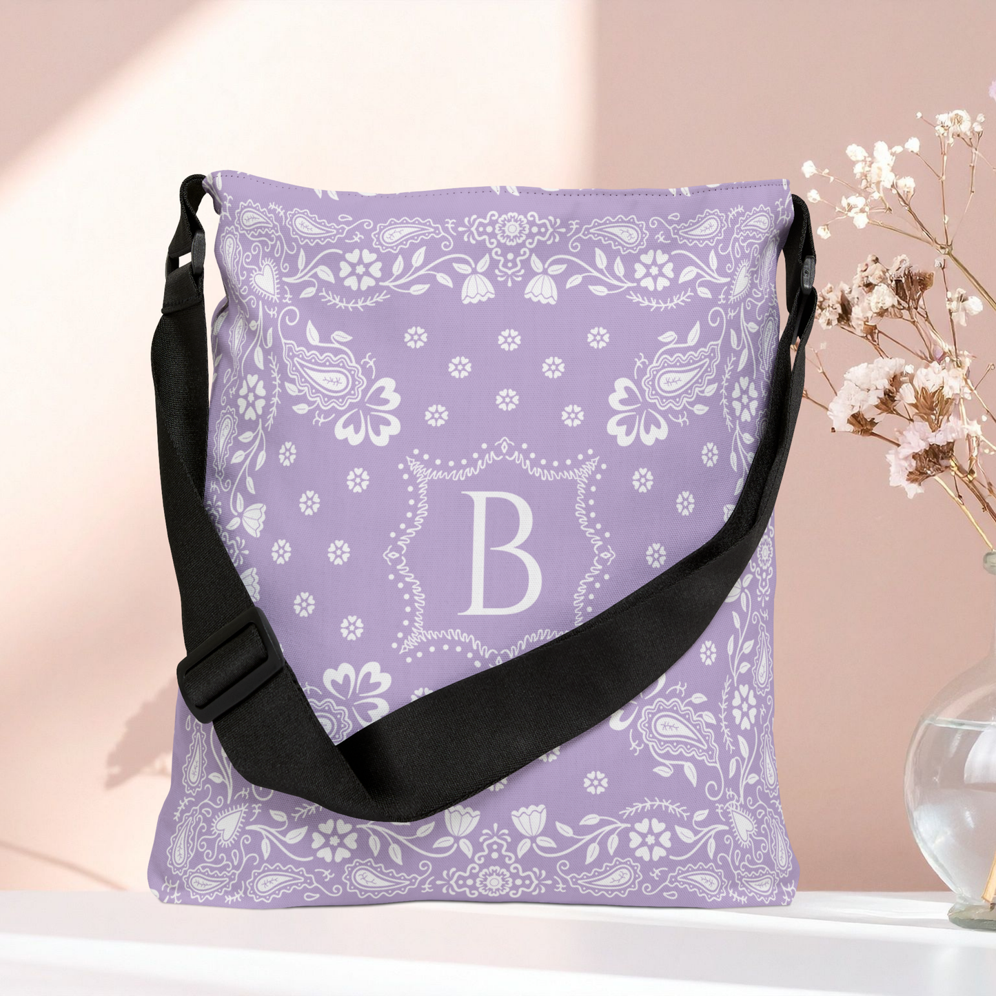 A lavender and white bandana patterned shoulder bag with a black strap, featuring a large monogram letter 'B' in the center, displayed on a white surface against a soft pink background with dried flowers in a vase.