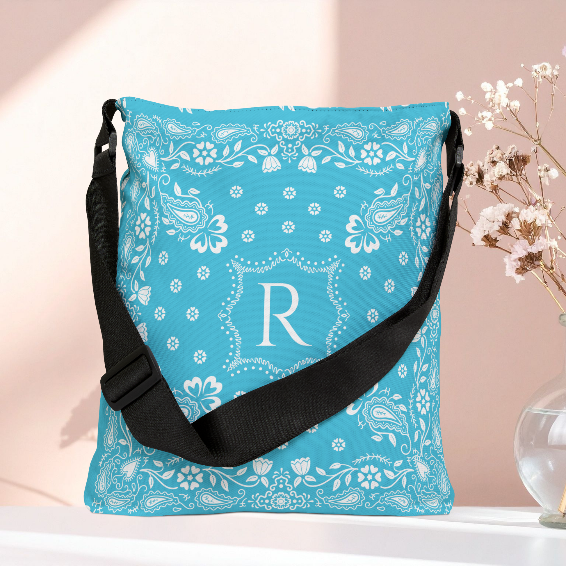 A turquoise blue shoulder bag with a white paisley bandana pattern and a large white monogram letter 'R' on the front, featuring a black adjustable strap. It is displayed on a white surface against a soft pink background with dried flowers in a glass vase on the right.