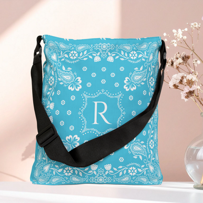 A turquoise blue shoulder bag with a white paisley bandana pattern and a large white monogram letter 'R' on the front, featuring a black adjustable strap. It is displayed on a white surface against a soft pink background with dried flowers in a glass vase on the right.