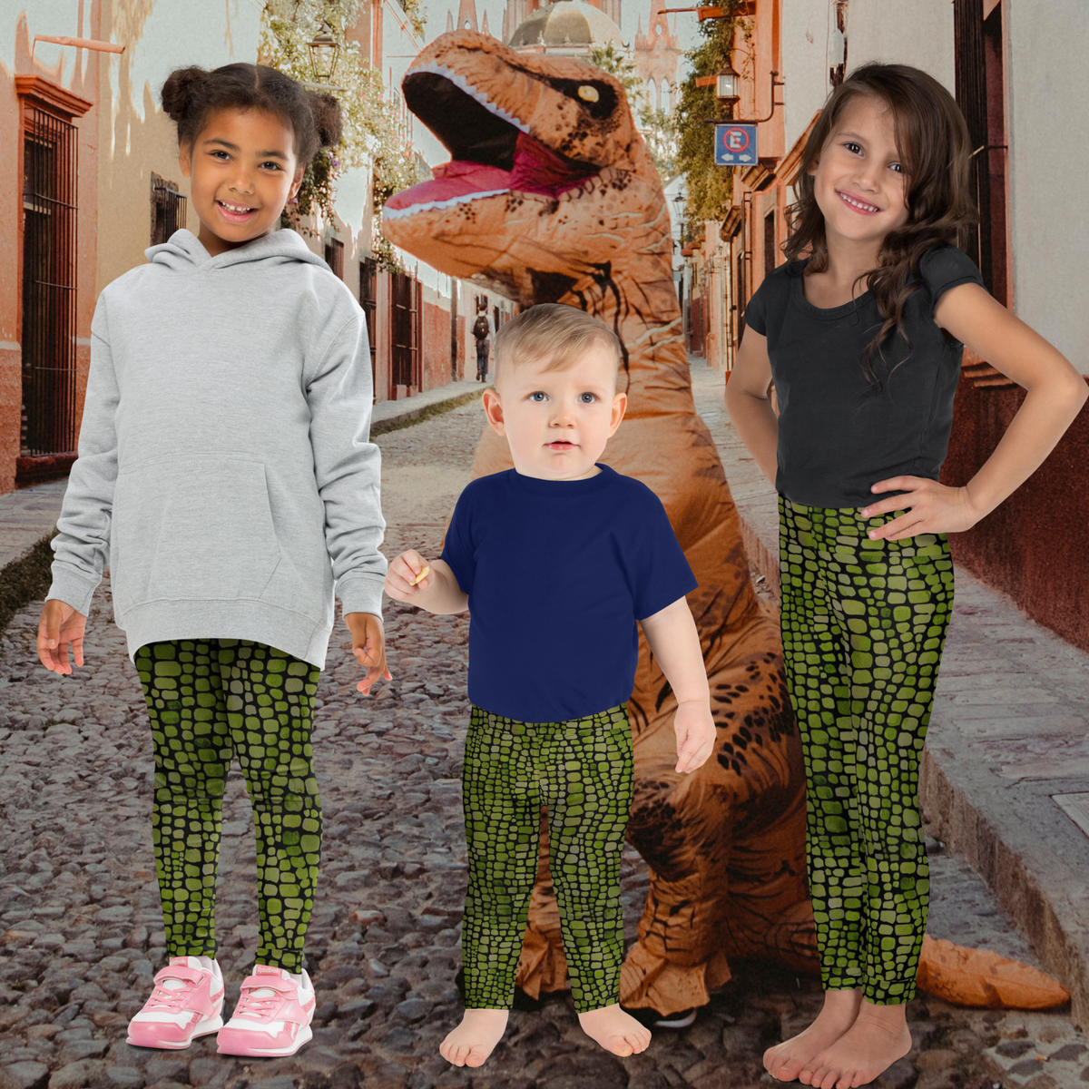 Two girls and a toddler boy wearing matching green and black reptile scale leggings stand beside a person in an inflatable T Rex costume. The kids show how these full length reptile print leggings can be used for dinosaur and dragon costumes, Halloween dress up, imaginative play, or everyday outfits. Available in sizes 2T through 20 for toddlers, kids, tweens, and teens.