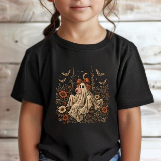 Toddler girl wearing a black Halloween T shirt featuring a whimsical vintage ghost blowing a bubble with bubble gum, surrounded by orange flowers, botanical foliage, bats, stars, lace details and a bow. A cute spooky season shirt for toddlers who love playful Halloween and fall outfits.