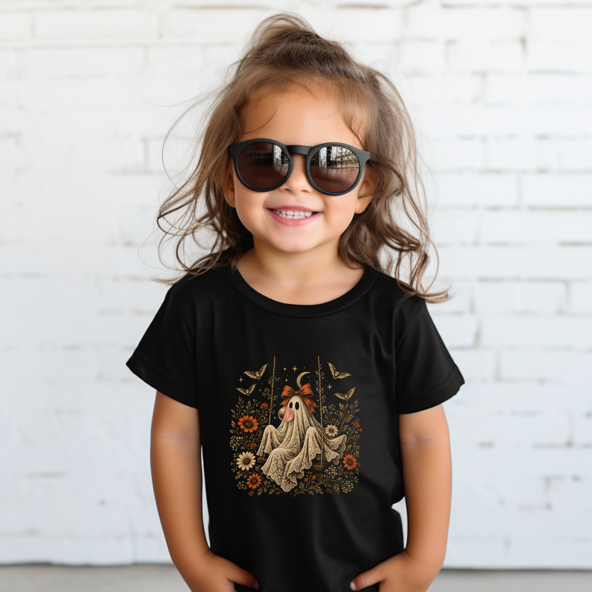 Toddler girl wearing a black Halloween T shirt with a whimsical ghost blowing bubble gum, surrounded by orange flowers, botanical foliage, bats, stars, lace and a bow. A vintage inspired spooky season shirt for toddlers, perfect for fall activities, Halloween celebrations and everyday wear.

