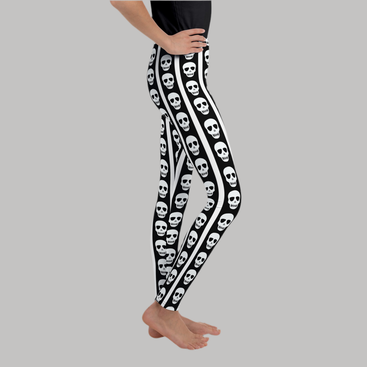 Black and white skull youth leggings featuring vertical stripe design with repeating skull pattern. Trendy spooky season leggings for girls and tweens sizes 8 to 20. Soft stretchy full length pants designed for comfort and movement. Perfect tween Halloween outfit, school fall wear, or casual everyday edgy fashion. Gothic inspired kids leggings ideal for trick or treat outfits, costume parties, and spooky themed styling.