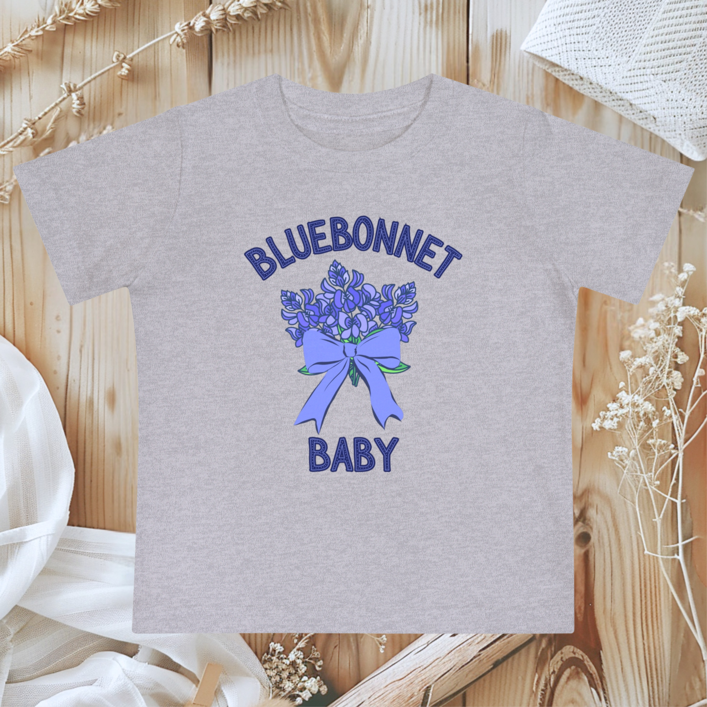 Athletic Heather Bella+Canvas 100B Bluebonnet Baby T-Shirt shown flat, highlighting its soft gray tone and delicate watercolor bluebonnet design inspired by Texas wildflowers. This breathable cotton baby tee offers comfort and southern charm for newborns to 24 months. A sweet Texas baby gift or spring wardrobe essential, it blends wildflower style with cozy everyday wear for nature-loving families.