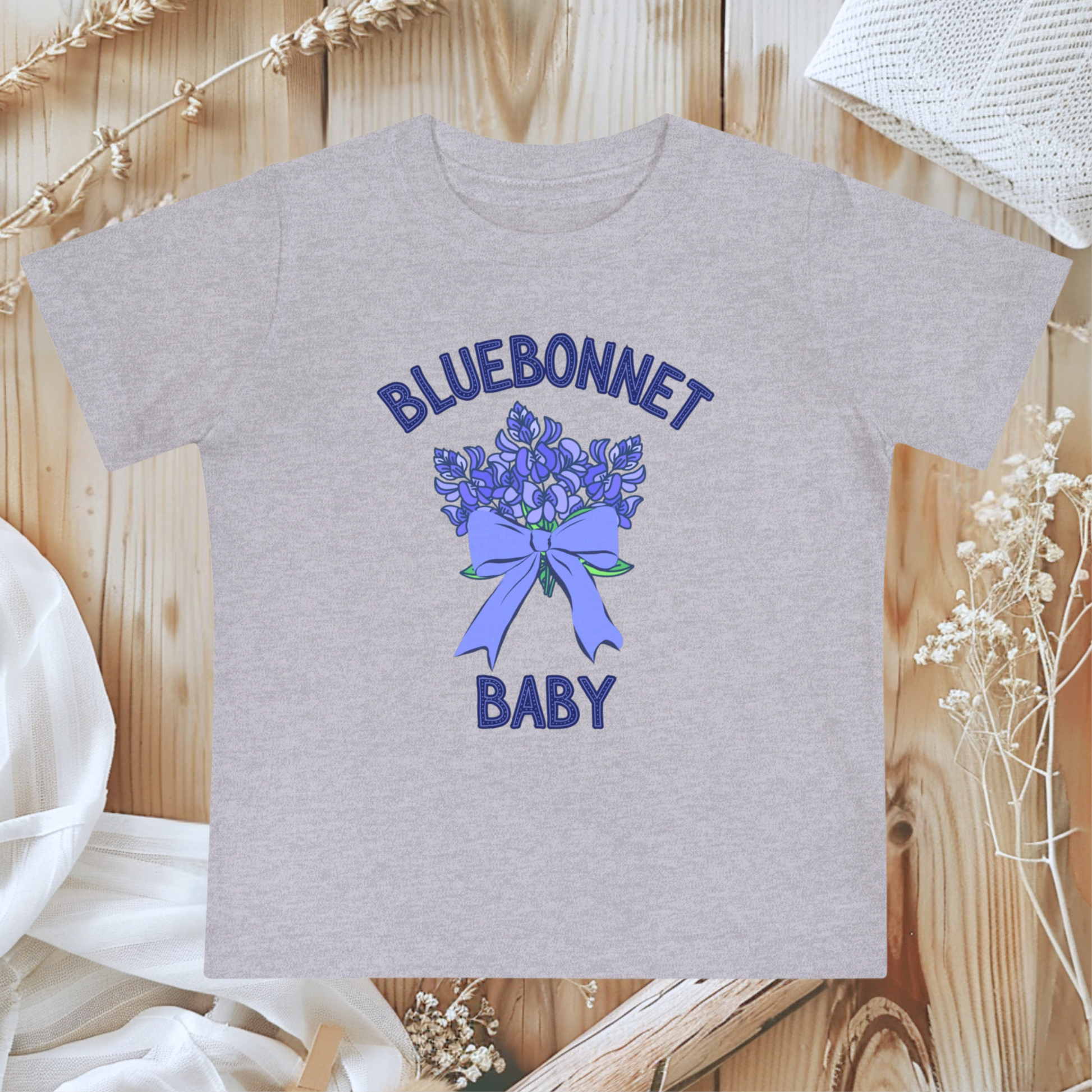 Athletic Heather Bella+Canvas 100B Bluebonnet Baby T-Shirt shown flat, highlighting its soft gray tone and delicate watercolor bluebonnet design inspired by Texas wildflowers. This breathable cotton baby tee offers comfort and southern charm for newborns to 24 months. A sweet Texas baby gift or spring wardrobe essential, it blends wildflower style with cozy everyday wear for nature-loving families.