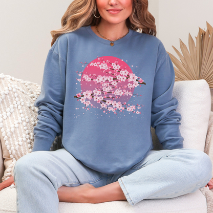 Blue Jean Comfort Colors sweatshirt styled as a soft spring casual outfit, featuring delicate pink sakura cherry blossoms and flowering branches across a dreamy pink moon graphic. The relaxed pullover combines a worn in vintage inspired feel with feminine floral artwork, making it an easy everyday layer for spring mornings, weekend outings, coffee runs, casual travel, and cherry blossom season.