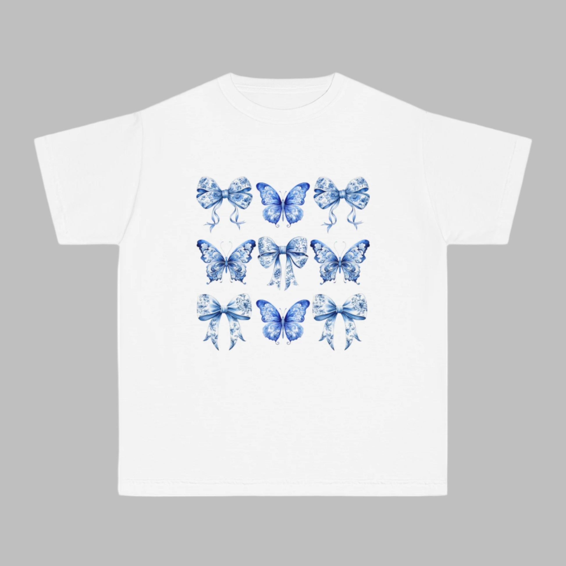 Image: Blue Toile Butterflies & Bows Girls T-shirt in soft Comfort Colors fabric, perfect for tweens and teens who love cottagecore, coquette fashion, or Texas-style charm. Featuring a dreamy blue toile print with vintage bows and delicate butterflies, this shirt adds classic southern flair to everyday outfits. Available in Chambray, White, True Navy, and Pepper, sizes XS–XL, part of a coordinating family matching collection, by jaecrece.