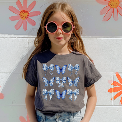 Image: Coquette-inspired girls Comfort Colors tee with a blue toile butterfly and bow print. This boutique-style youth shirt is perfect for girls who love trendy Texas aesthetics and vintage countryside patterns. Sizes XS–XL in Chambray, White, True Navy, and Pepper. Part of a mommy and me matching outfit collection, perfect for gifting, events, or family photos, by jaecrece.