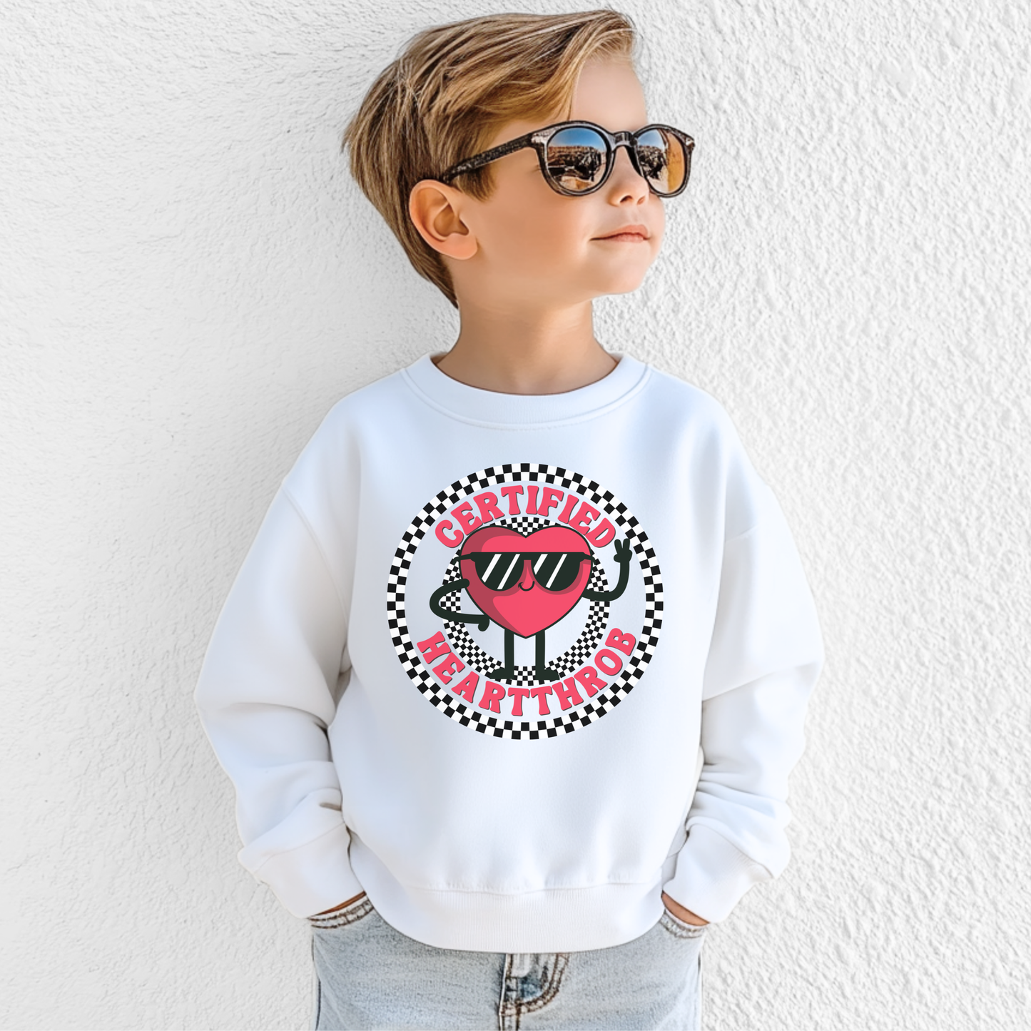 A child models a white Certified Heartthrob youth sweatshirt featuring a playful cartoon heart character wearing sunglasses. The heart is surrounded by a bold black and white checkerboard circle, with Certified Heartthrob lettering around the design. The fun graphic has a retro inspired feel that makes this sweatshirt a cheerful choice for Valentine's Day.