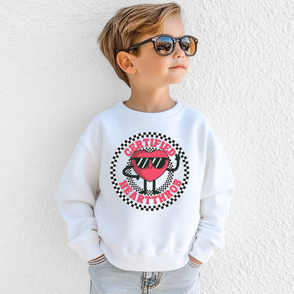 A child models a white Certified Heartthrob youth sweatshirt featuring a playful cartoon heart character wearing sunglasses. The heart is surrounded by a bold black and white checkerboard circle, with Certified Heartthrob lettering around the design. The fun graphic has a retro inspired feel that makes this sweatshirt a cheerful choice for Valentine's Day.