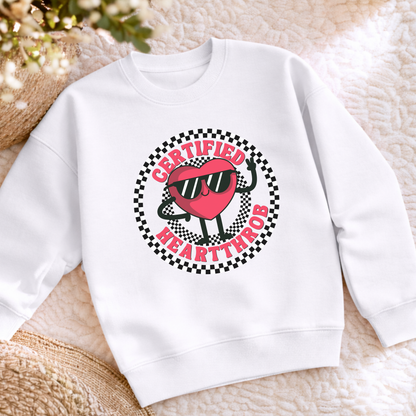 A white youth sweatshirt decorated with a playful Certified Heartthrob Valentine's Day design. A cartoon heart character wearing sunglasses sits inside a bold black and white checkerboard circle, with the phrase Certified Heartthrob surrounding the illustration. The cheerful graphic adds personality to a child's winter wardrobe and makes a fun seasonal gift.